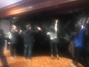 Four adults are lined along and facing a curving wall covered in black synthetic fur. They stand on a wooden floor with backs facing outward as the each touch the wall moving both hands along and around the wall's surface.